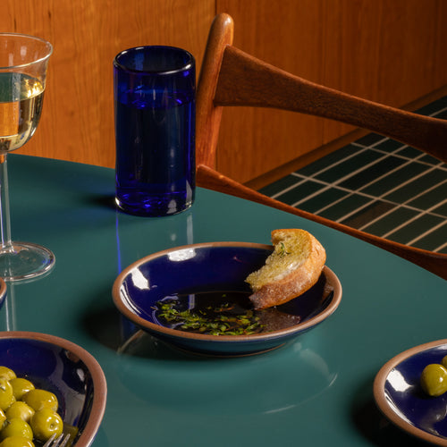 Glossy dark blue ceramic plate with a bowl edge with olive oil, herbs, and a piece of bread on a green table.