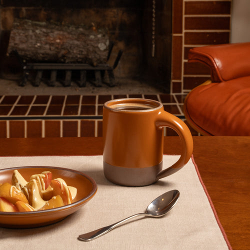Earthy orange ceramic mug and plate with a dessert and spoon on a linen tablecloth in front of a fireplace.