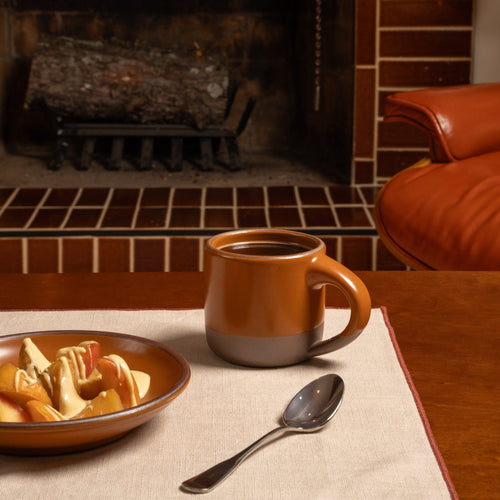 A small warm ceramic mug with a earth  orange upper and brown clay lower band sits on a table in a cozy room with a brick fireplace. A matching bowl of fruit and a spoon are nearby on a beige placemat.