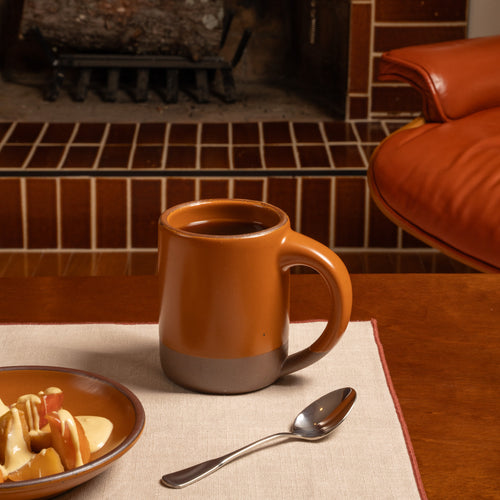 A ceramic mug with a earthy orange upper body and a gray lower band sits on a table in a cozy indoor setting with a fireplace, a spoon, and a plate of fruit nearby.
