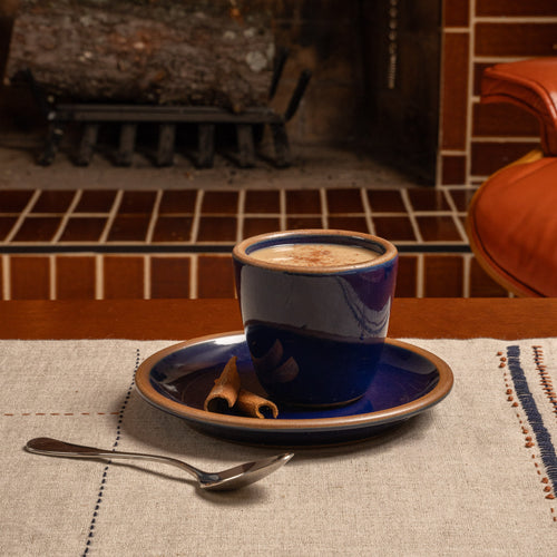 A glossy dark blue ceramic cup and plate with a creamy drink and cinnamon sticks on a linen tablecloth.