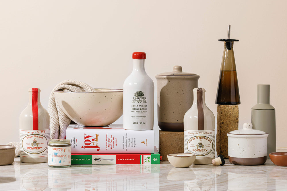 A styled product arrangement of kitchen tools and ingredients against a warm beige background, including ceramic bowls, bottles, jars, books, and a small towel.