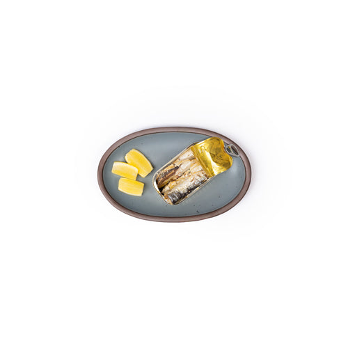 A small oval ceramic platter in a blue-grey color featuring iron speckles and an unglazed rim, with sardines