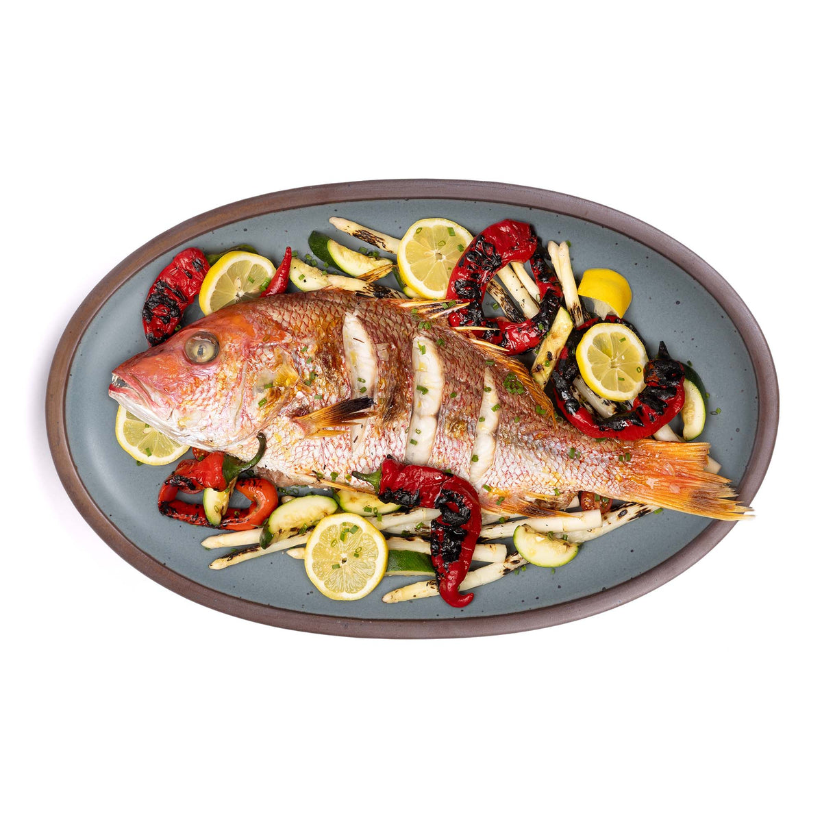 A large oval ceramic platter in a blue-grey color featuring iron speckles and an unglazed rim, with a whole fish along with lemons and more.