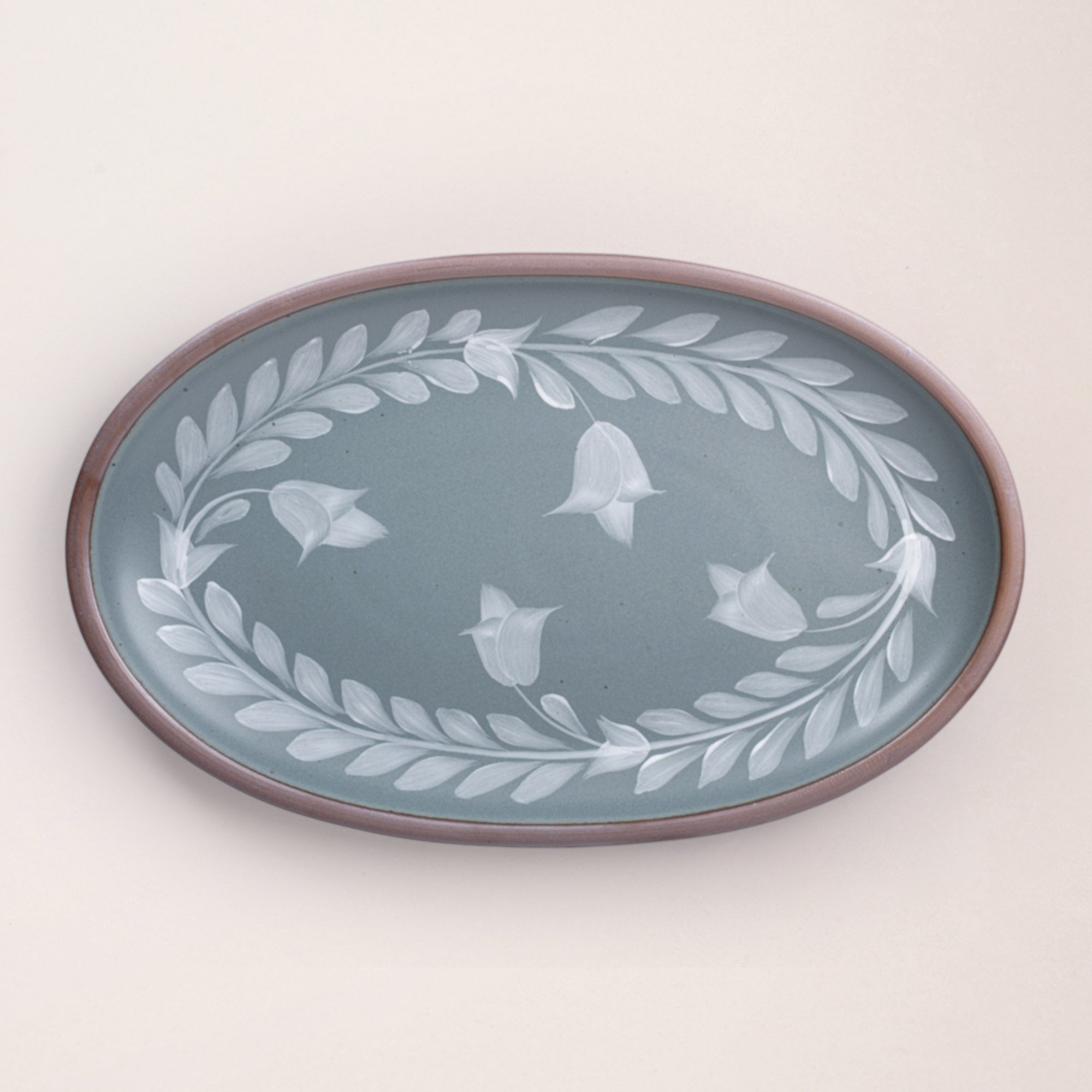 Oval ceramic platter with a hand-painted white leaf and flower border on a blue-grey background with an exposed clay rim.