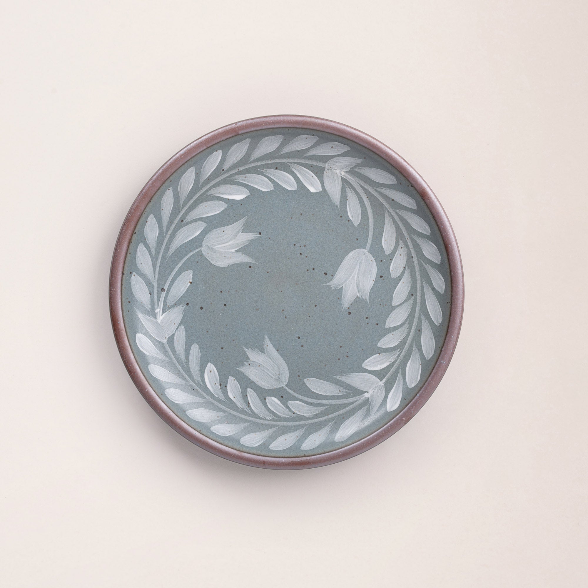Dinner round ceramic plate with a hand-painted white leaf wreath and floral design on a blue-grey background with an exposed clay rim.