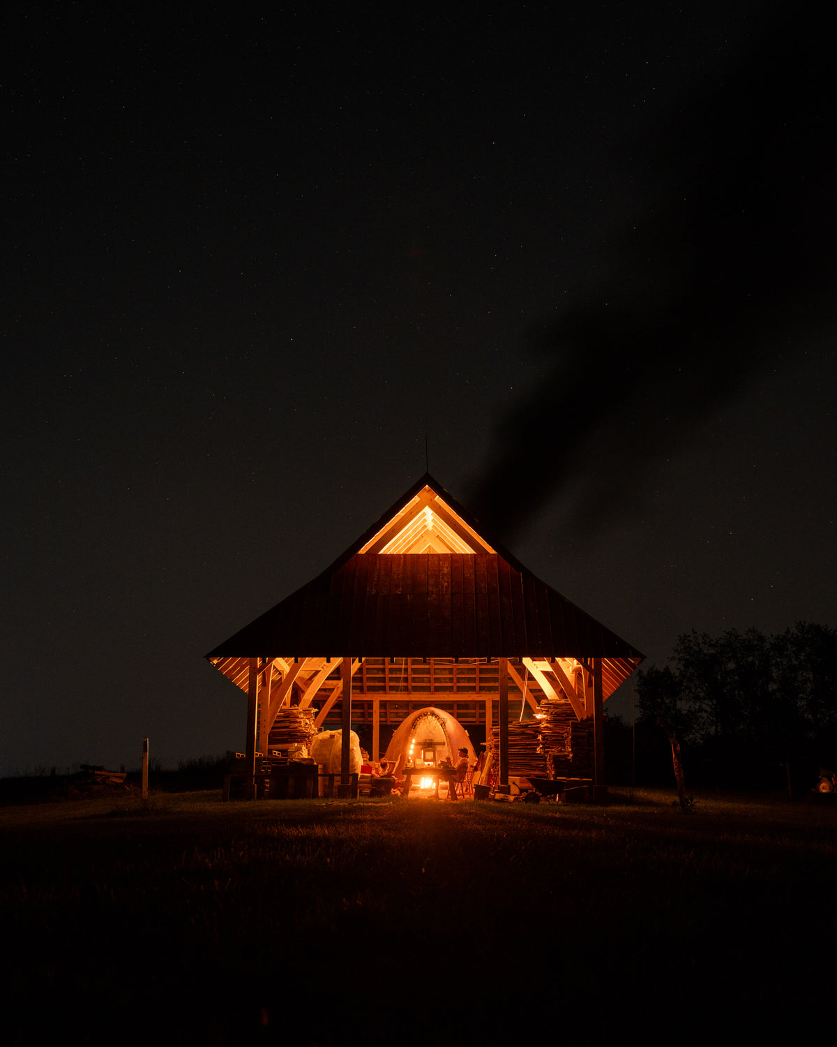 A cabin at night with a wood fired kiln firing with the whole cabin aglow and smoke coming out of the top.