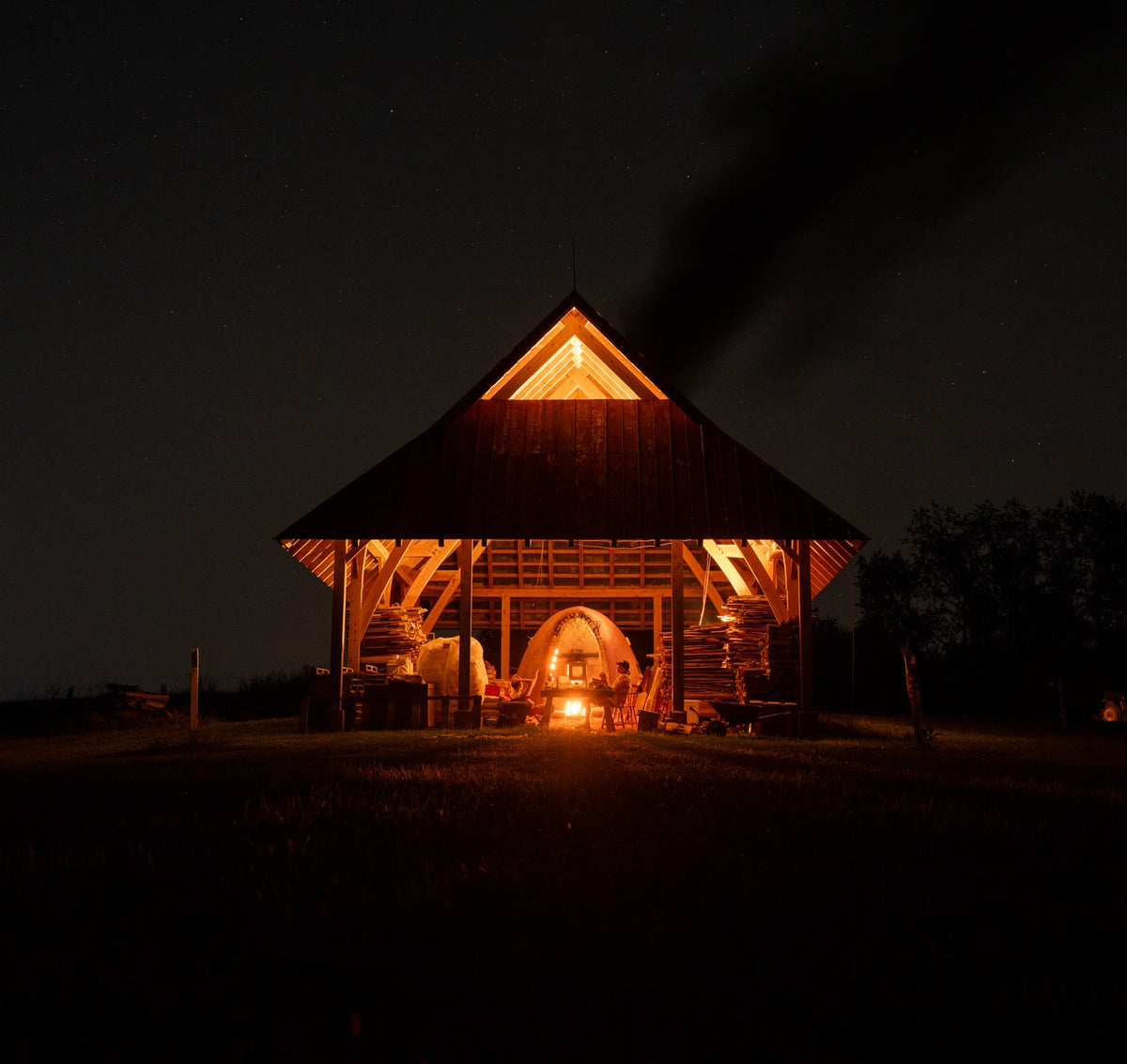 A cabin at night with a wood fired kiln firing with the whole cabin aglow and smoke coming out of the top.