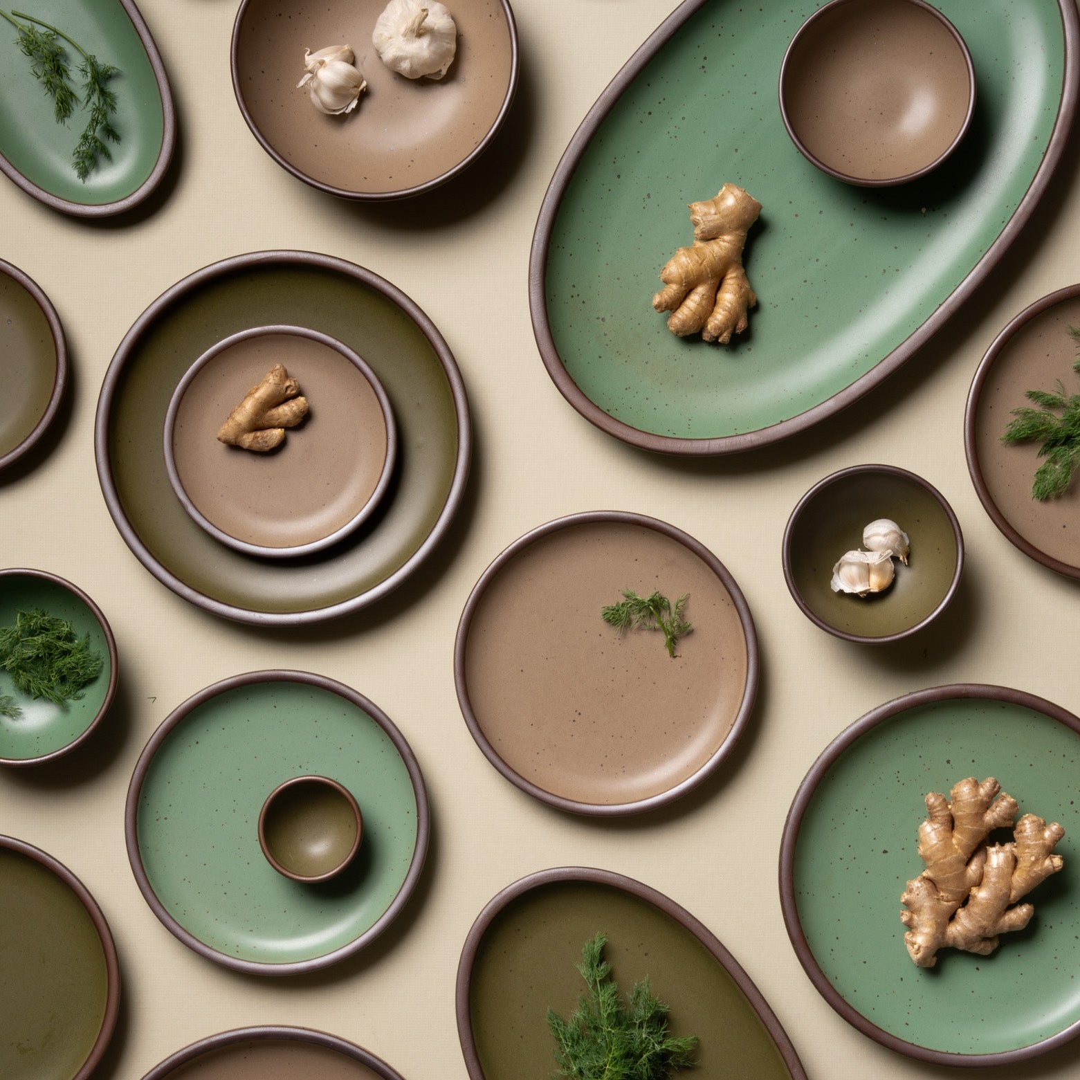 A styled flat-lay arrangement of ceramic tableware in earthy tones, including plates, bowls, and serving dishes in sage green, taupe, and brown, decorated with ginger, garlic, and herbs on a beige background.