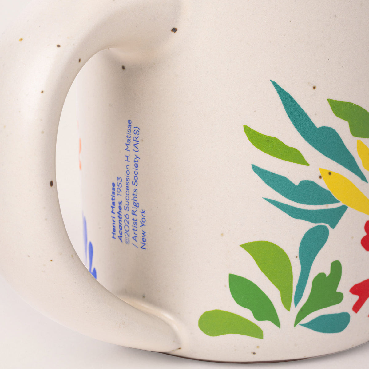 Close-up of a ceramic mug handle and side showing blue printed artist text and a colorful floral pattern in green, teal, yellow, and red on an off-white speckled glaze.