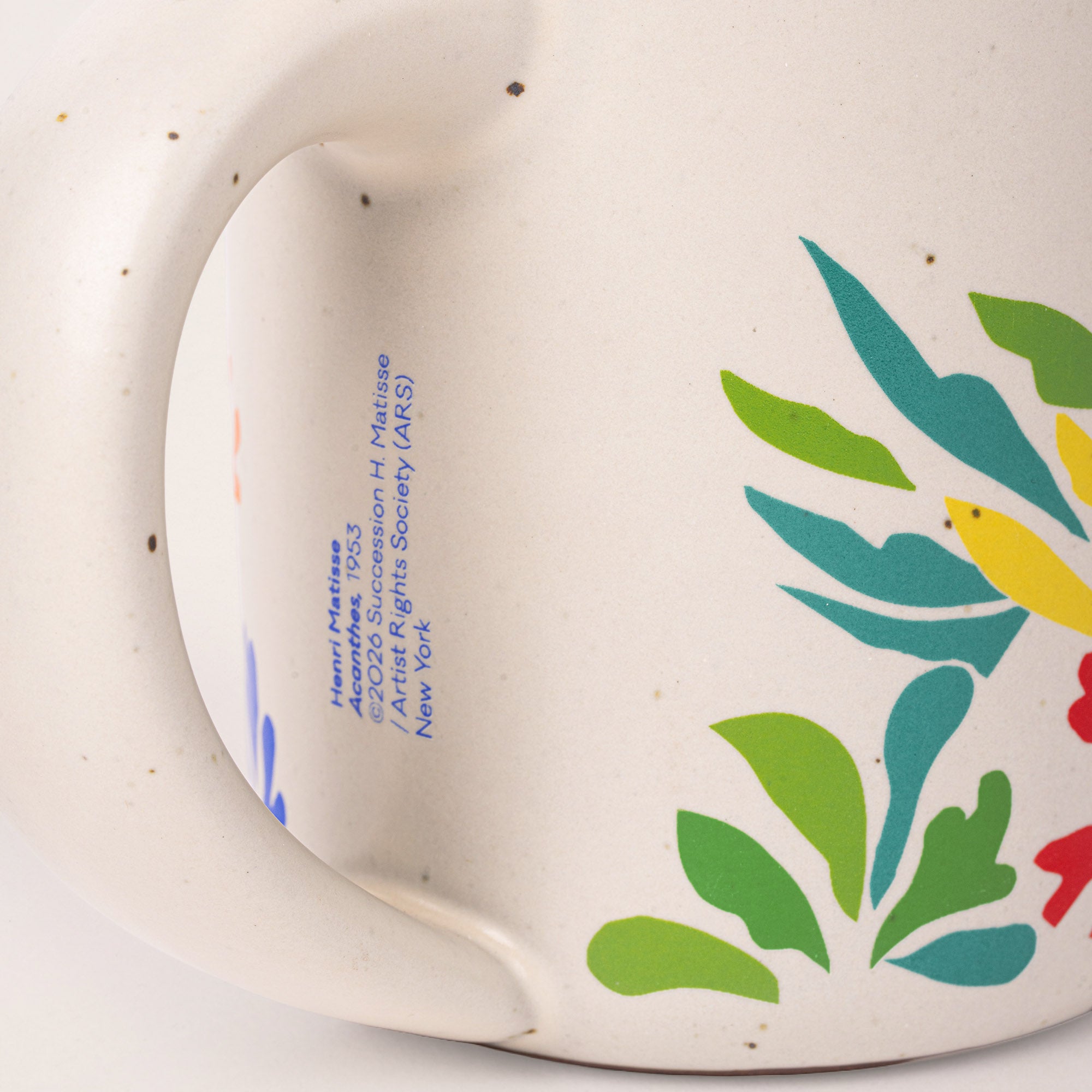 Close-up of a ceramic mug handle and side showing blue printed artist text and a colorful floral pattern in green, teal, yellow, and red on an off-white speckled glaze.