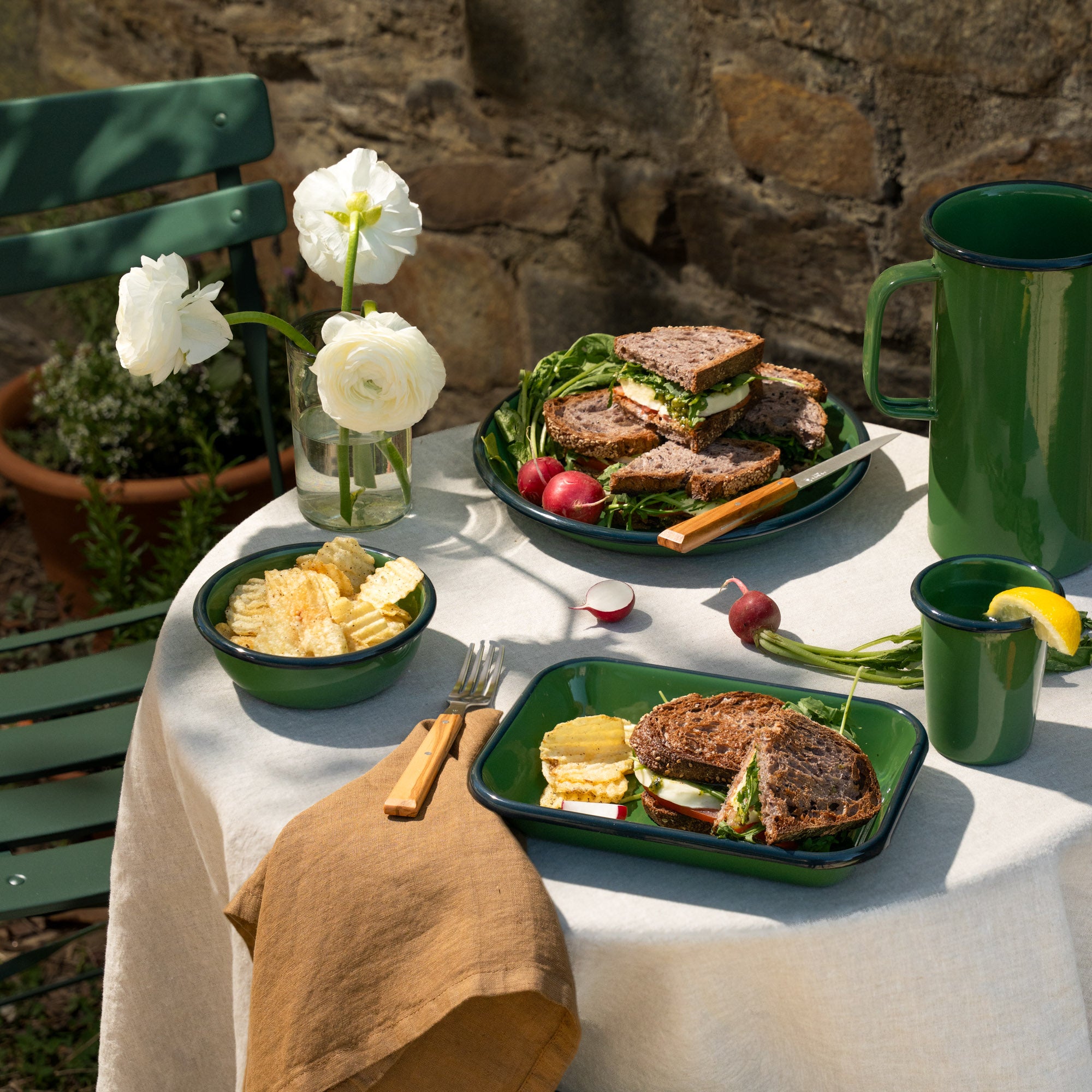 Outdoor table set with green enamelware, sandwiches, chips, radishes, and drinks, with white flowers in a vase beside a stone wall.