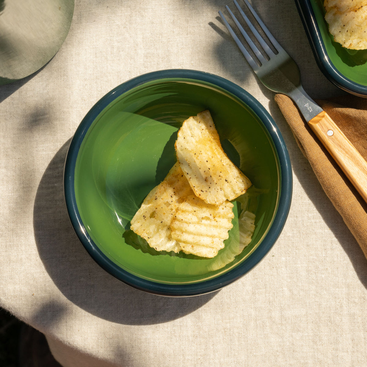 A small green enamel bowl with a dark rim, containing two ridged potato chips, styled on a beige linen tablecloth with a wooden-handled fork nearby.
