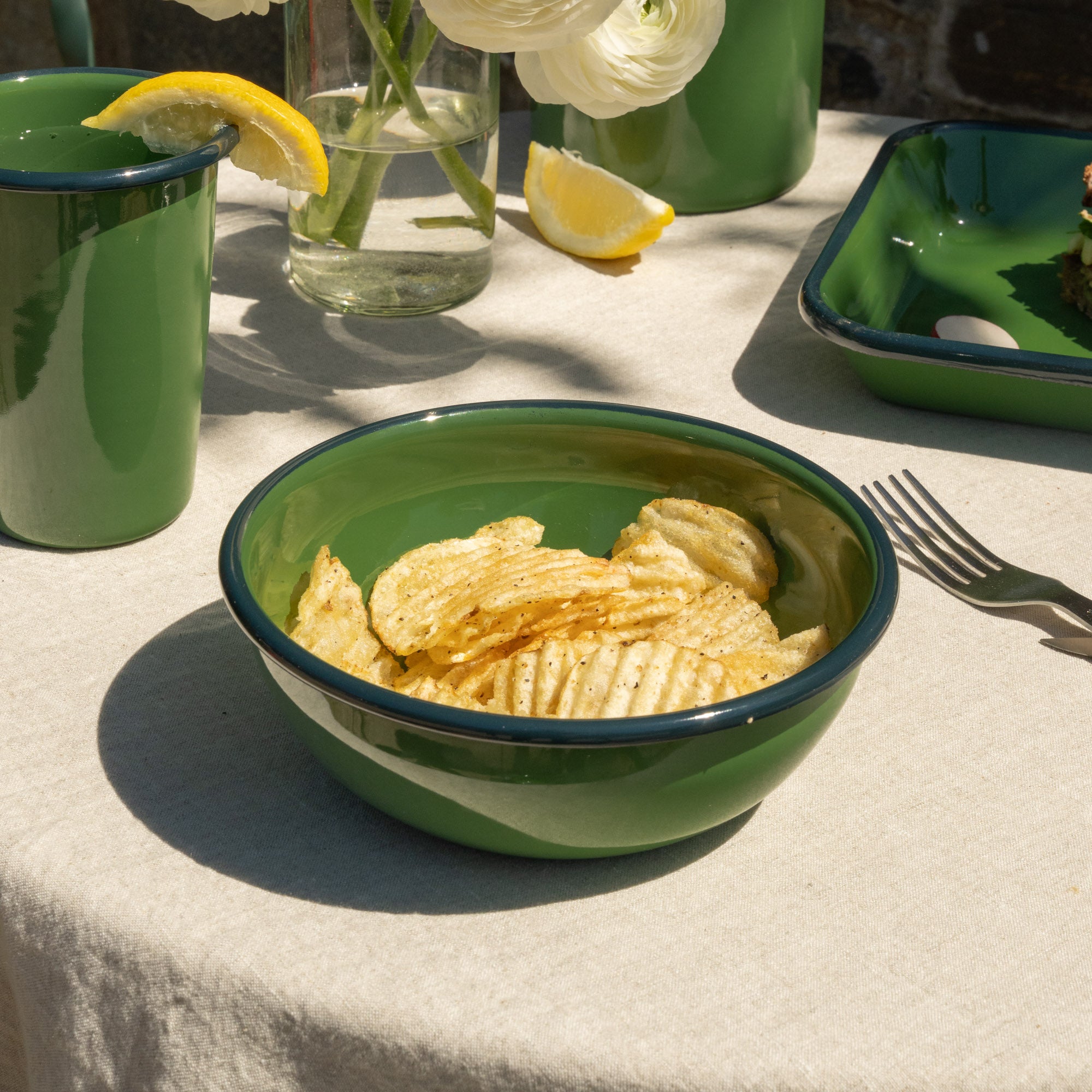 A medium green enamel bowl filled with ridged potato chips on a beige tablecloth, with matching green tableware and lemon slices in the background.
