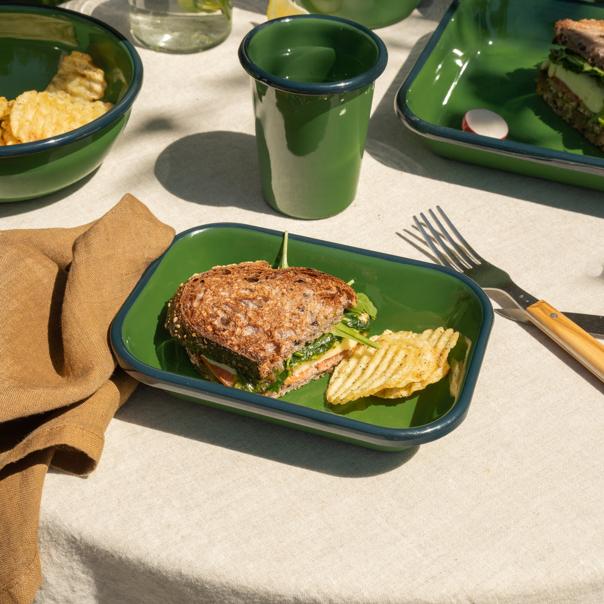 Small rectangular baking dish in a green color with a dark green rim, filled with a half sandwich and some chips on a tablecloth outside.