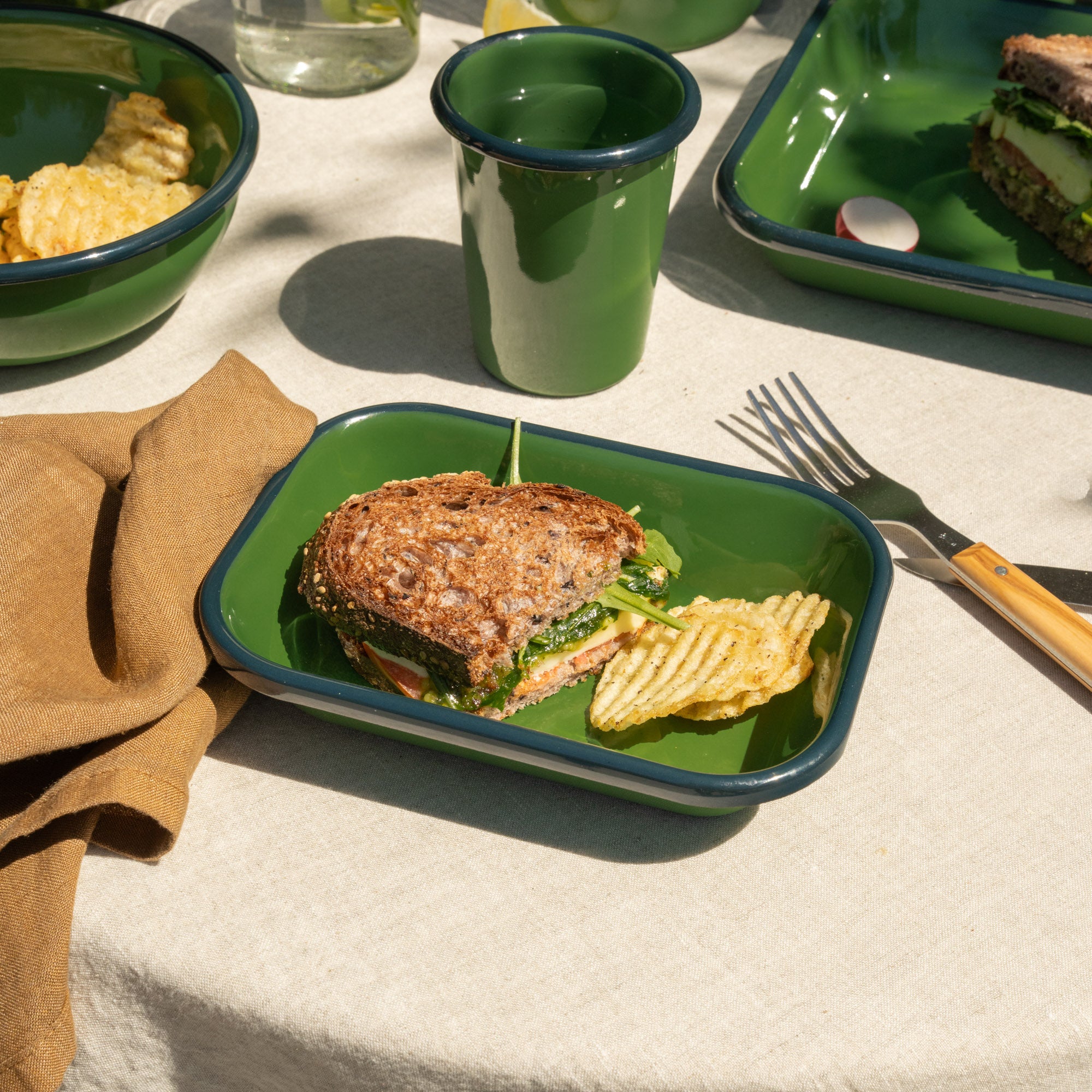 Small rectangular baking dish in a green color with a dark green rim, filled with a half sandwich and some chips on a tablecloth outside.