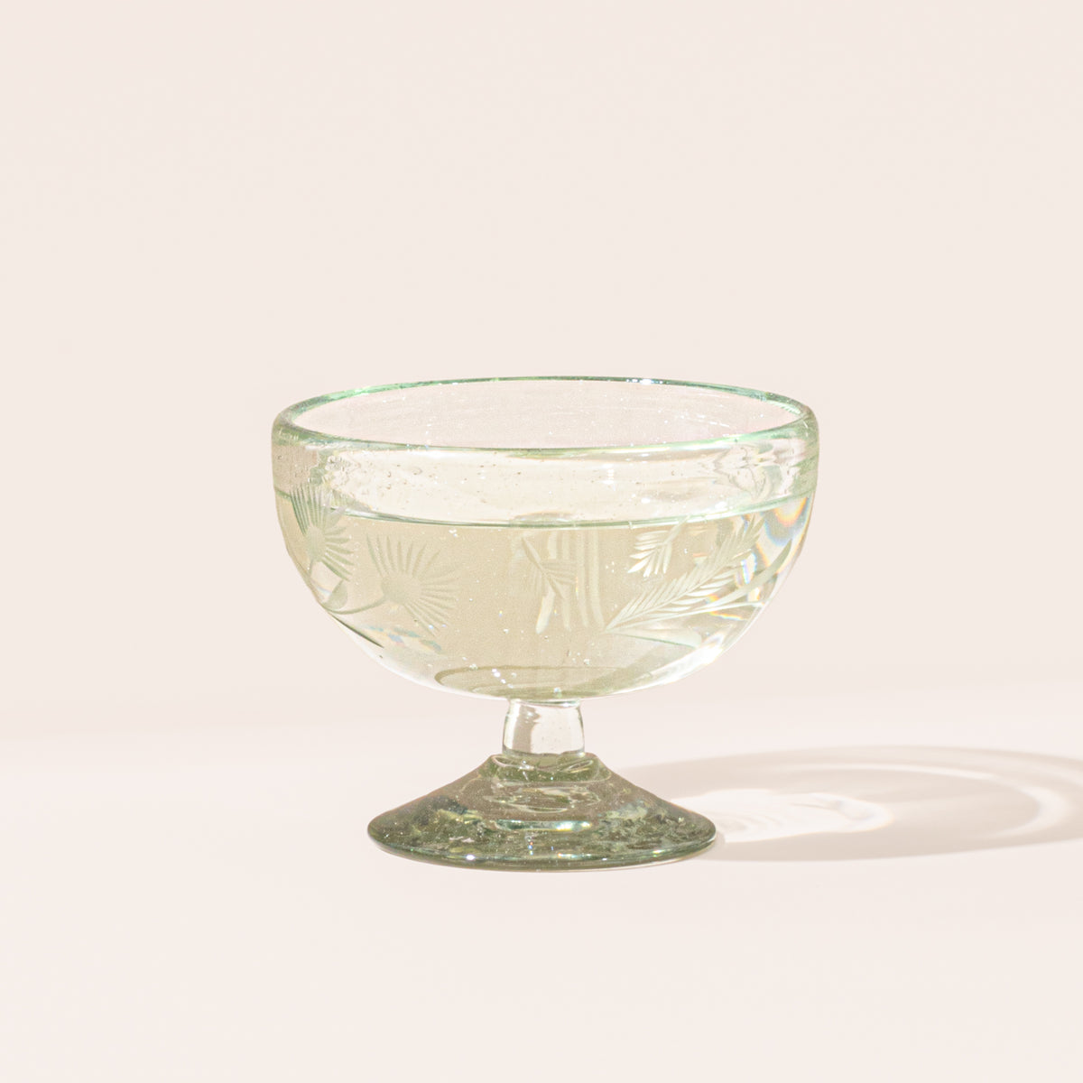 A clear etched coupe glass with a pedestal base and delicate botanical pattern with a drink inside.
