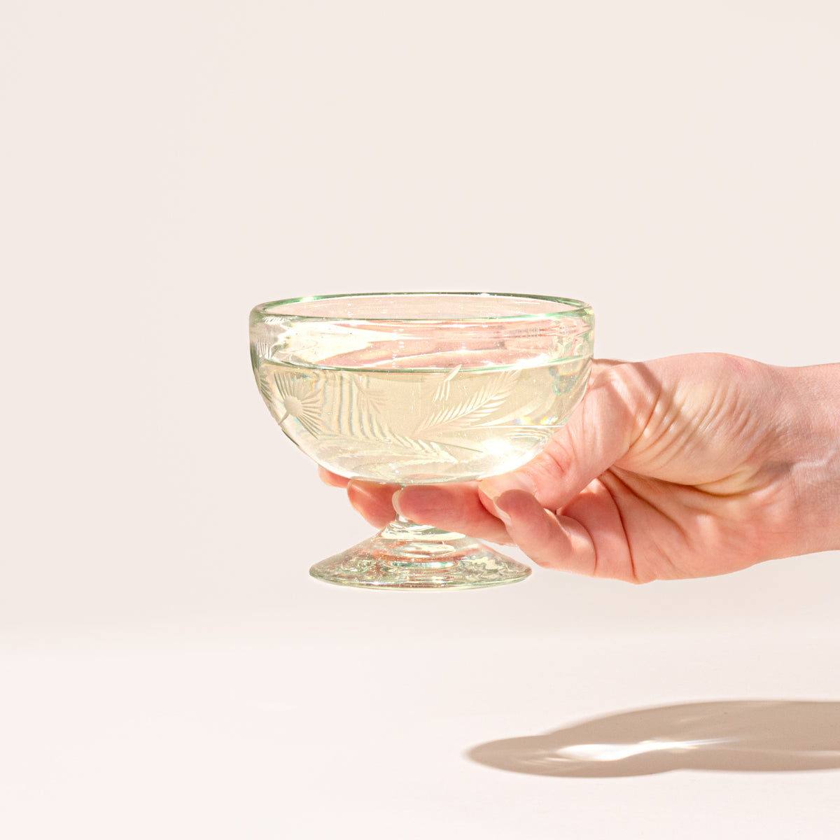 A hand holds a clear etched coupe glass with a pedestal base and delicate botanical pattern with a drink inside.