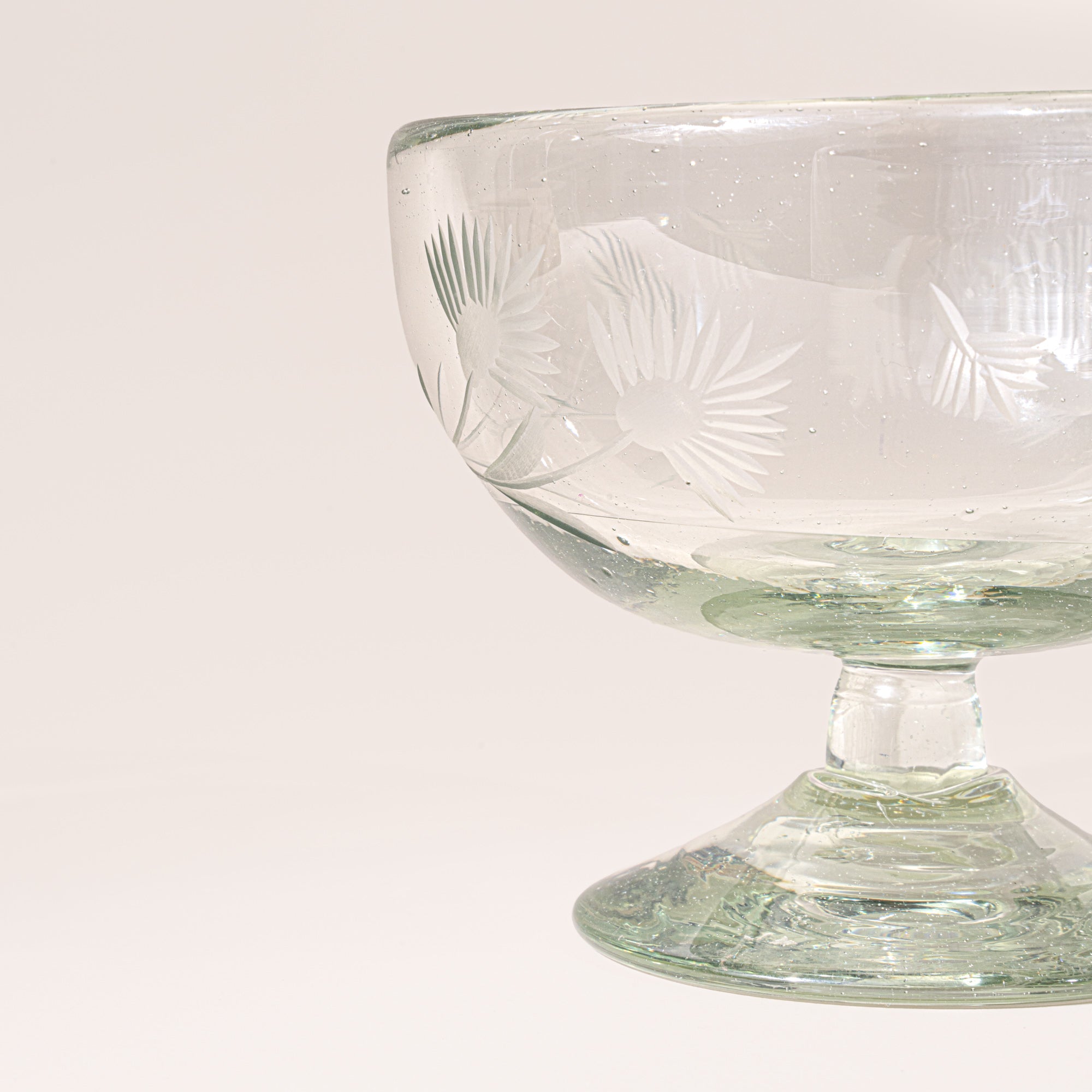 A closeup of a clear etched coupe glass with a pedestal base and delicate botanical pattern, shown on a clean neutral background.