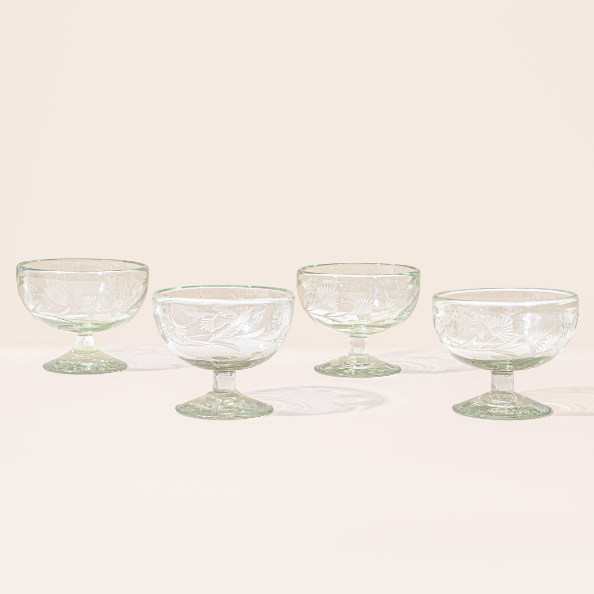 Four clear etched coupe glasses with a pedestal base and delicate botanical pattern, shown on a clean neutral background.