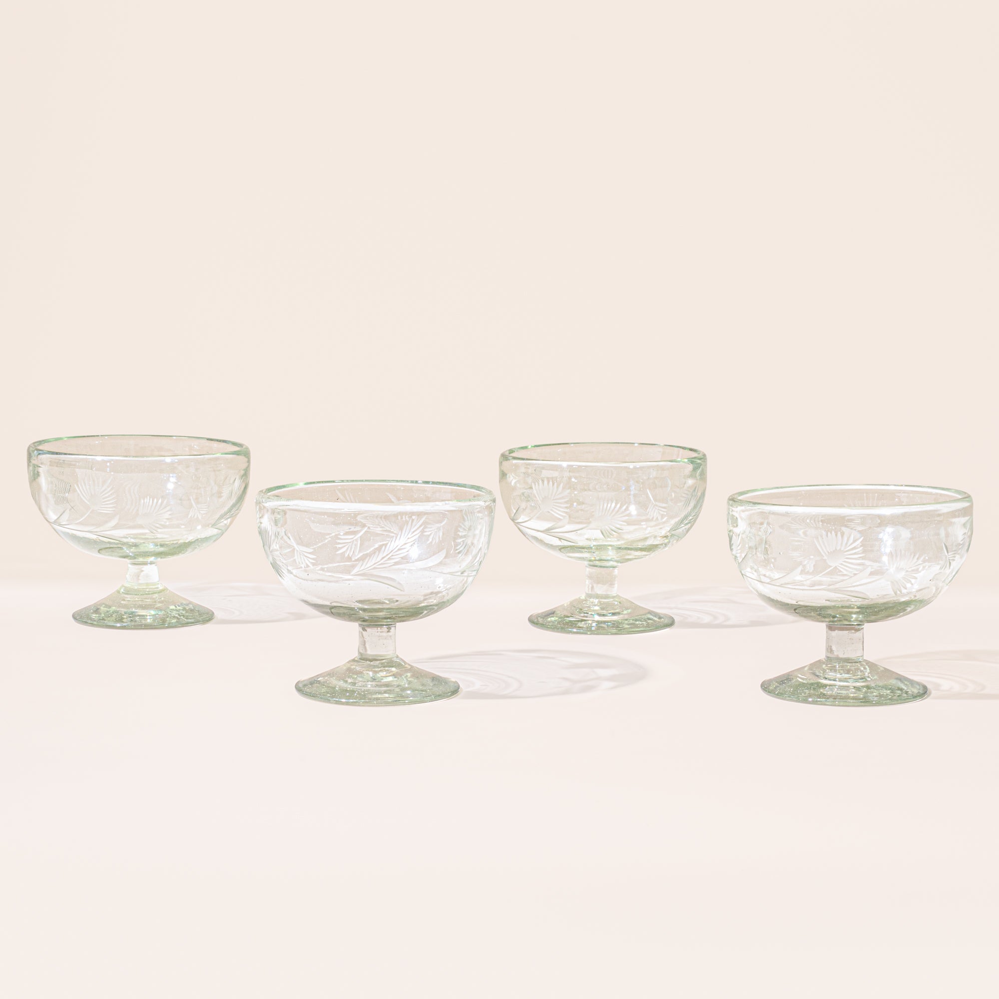 Four clear etched coupe glasses with a pedestal base and delicate botanical pattern, shown on a clean neutral background.