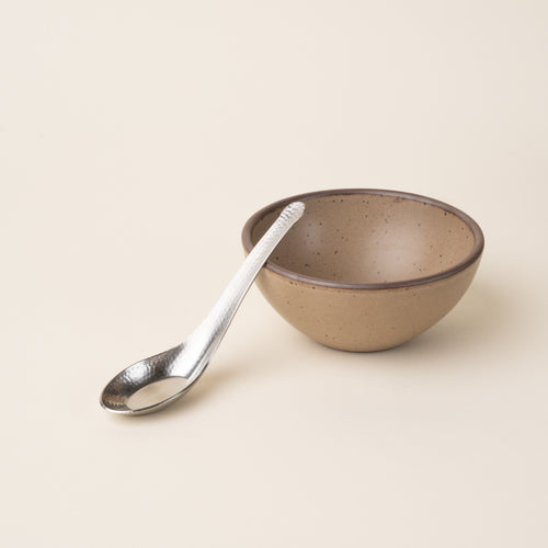A steel Asian soup spoon with hammered finish leans on a ceramic round bowl