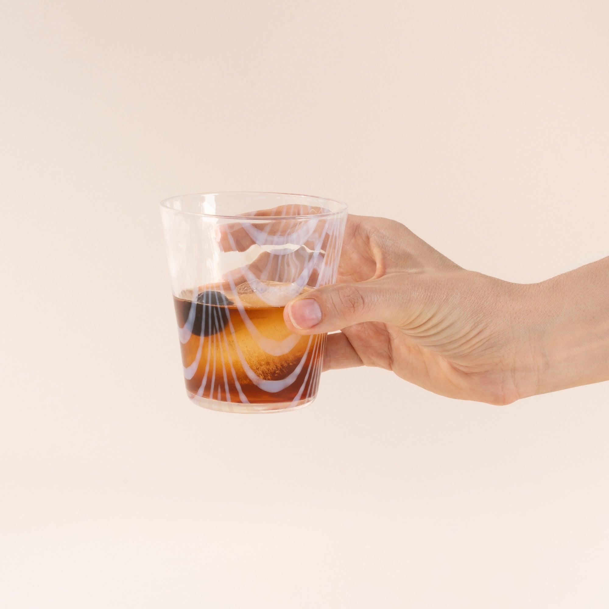 A hand holds a short clear glass with a delicate ripple pattern, half filled with a drink