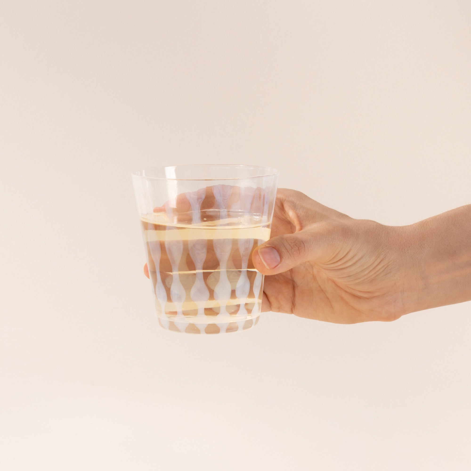A hand hold a short clear glass with a delicate vertical wave pattern, half filled with a drink
