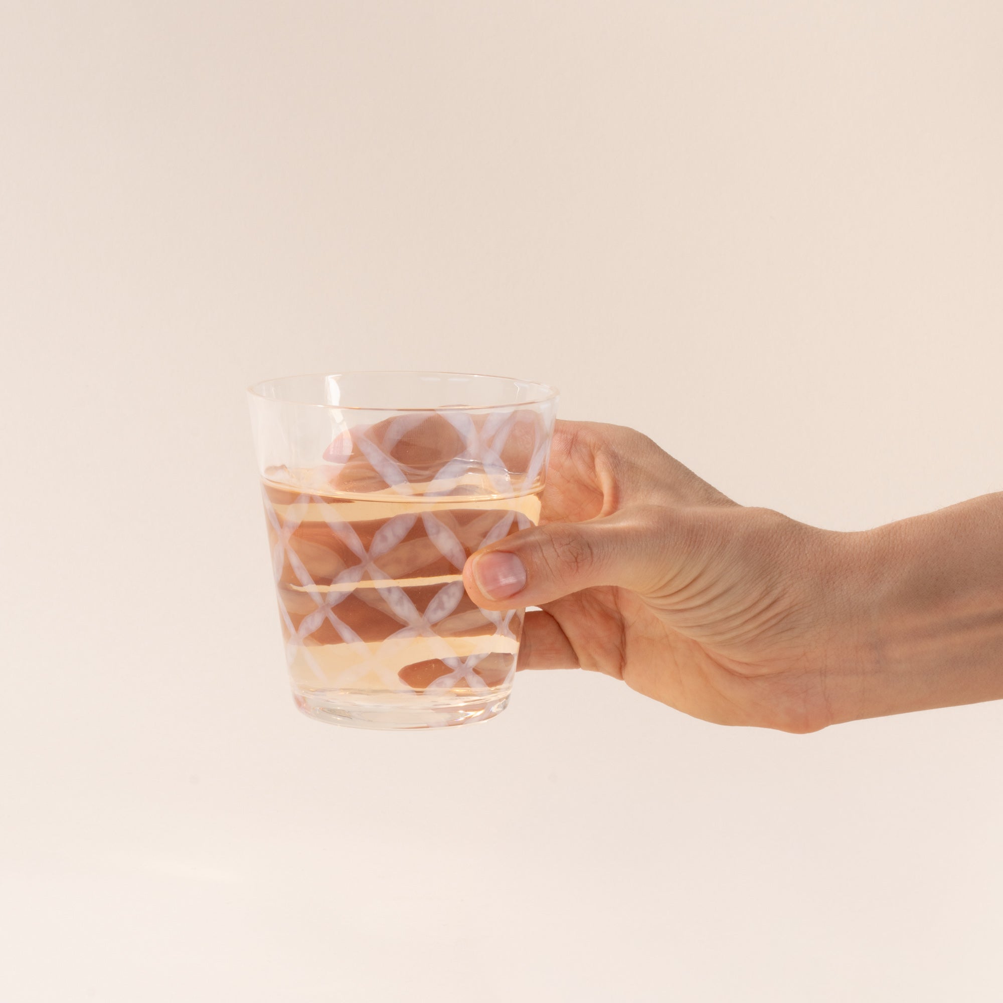 A hand holds a short clear glass with a delicate grid pattern, half filled with a drink