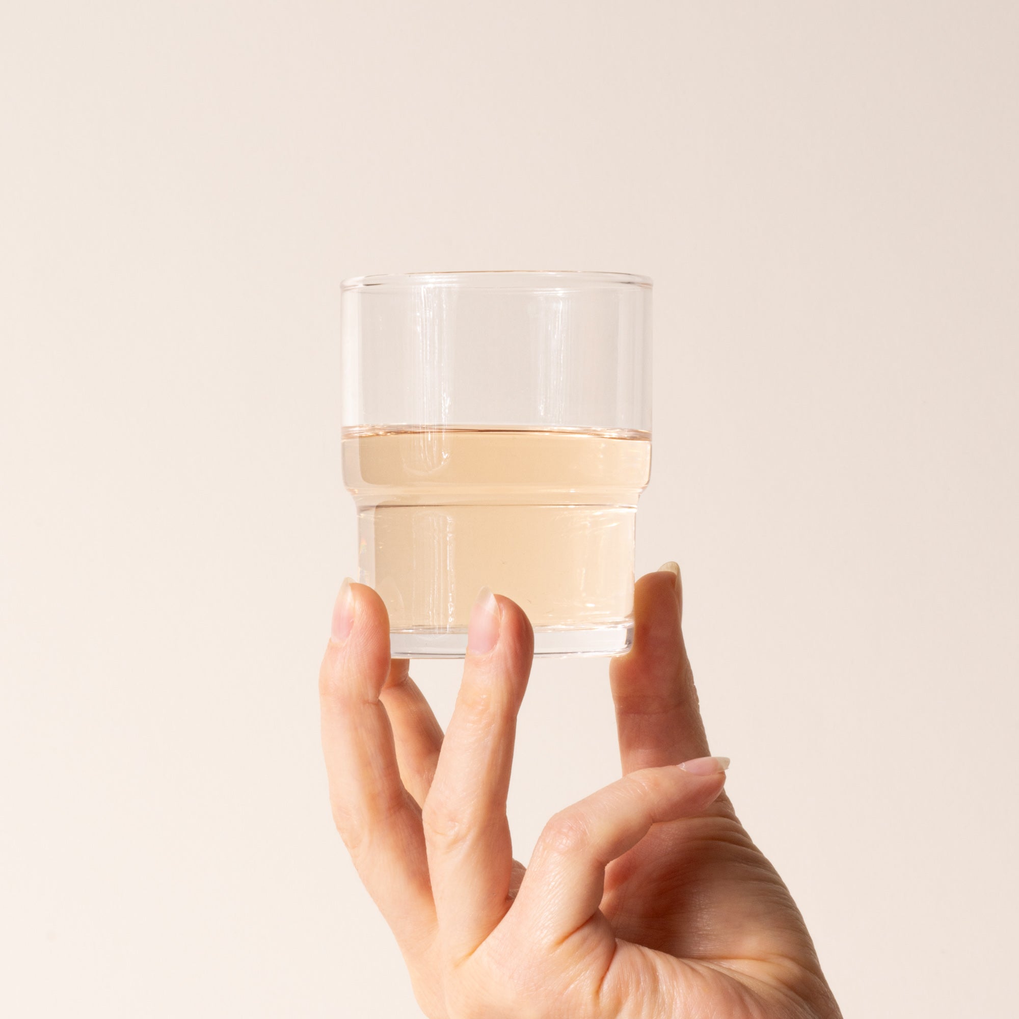 Hand holds a clear cylindrical glass with a wider top half with a narrower bottom half, filled with a drink.