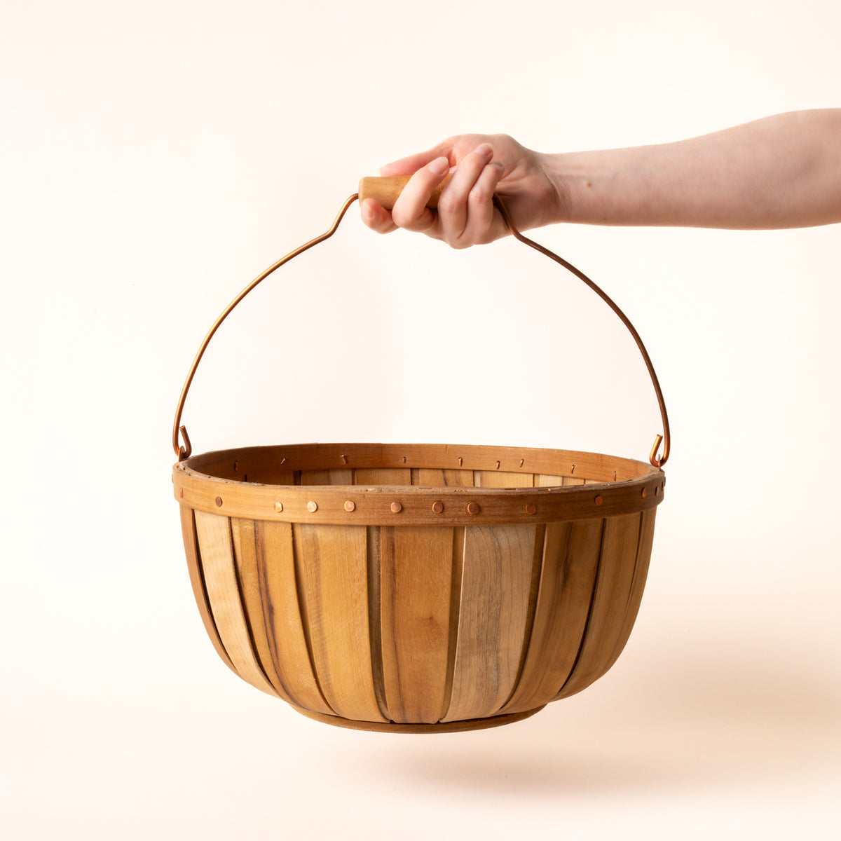 A hand holds a round wooden gathering large basket with copper handle and rivet details, shown against a light background.