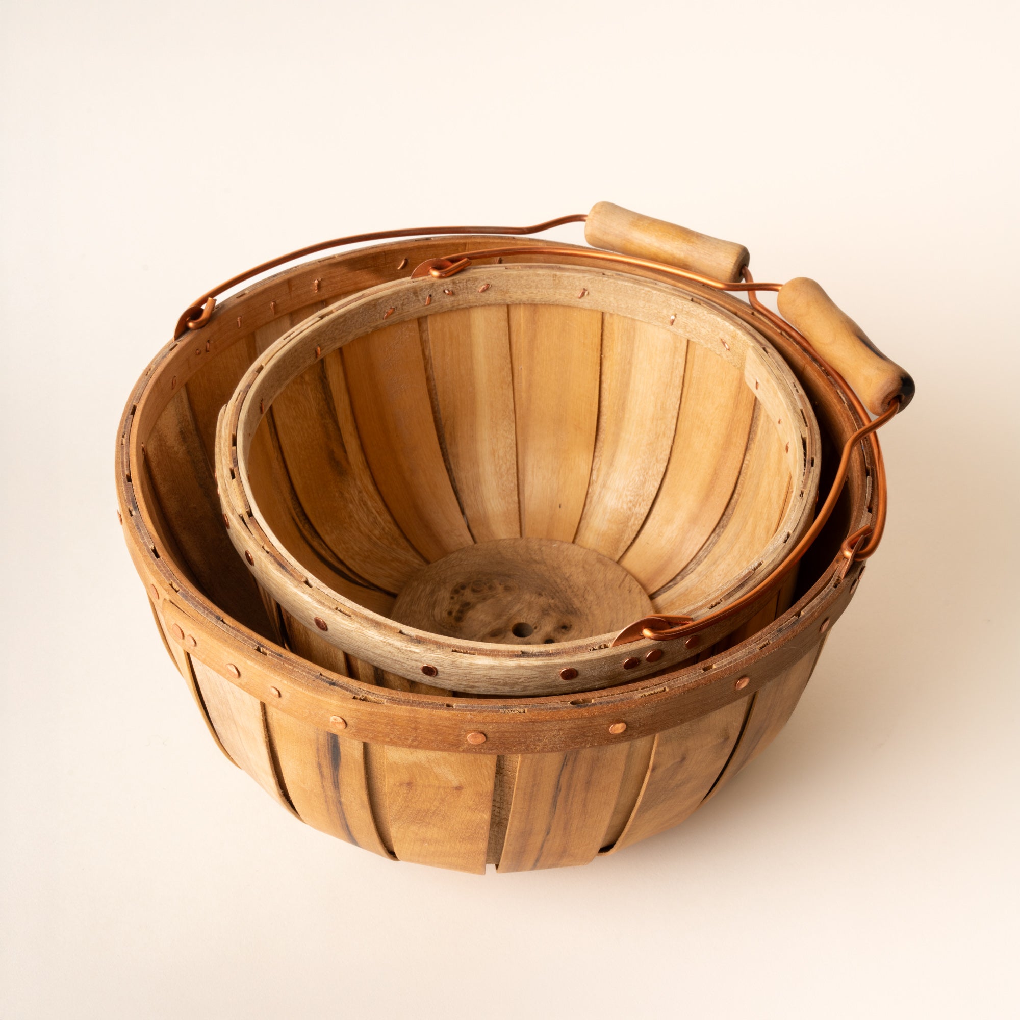 Two nested wooden bushel baskets with copper handles and rivets, showing slatted interiors and natural wood grain.