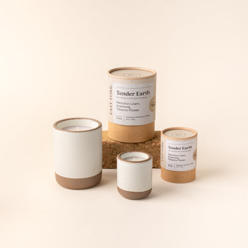 Small and large ceramic vessel in a cool white color with candles inside, arranged with tube packaging that reads 'Tender Earth'