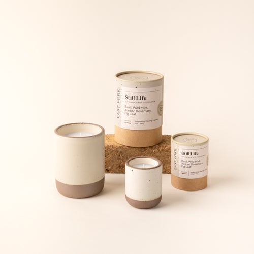 Small and large ceramic vessel in a warm off-white color with candles inside, arranged with tube packaging that reads 'Still Life'