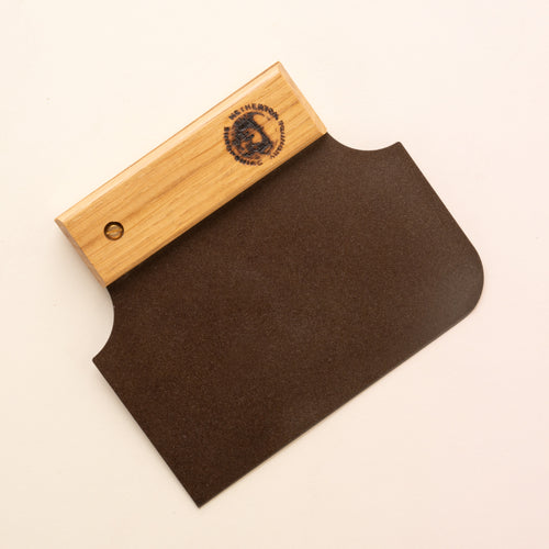 A black metal bench scraper with one rounded corner on the blade end while the handle end tapers in to a narrow wooden handle 