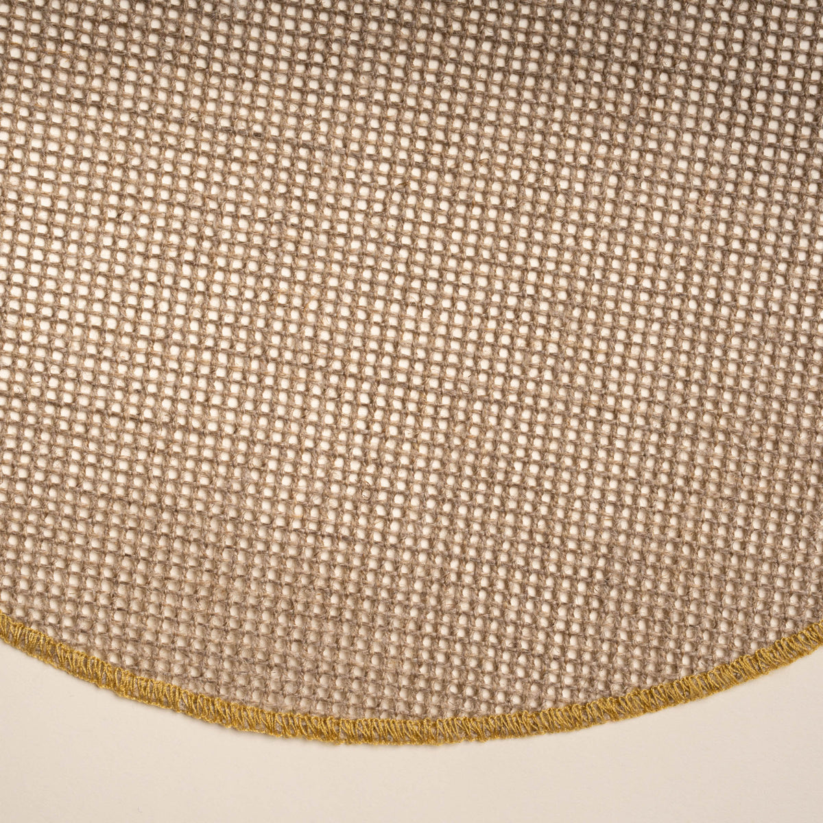 Closeup of a round woven placemat in a natural color with a sewn ochre rim on a beige background