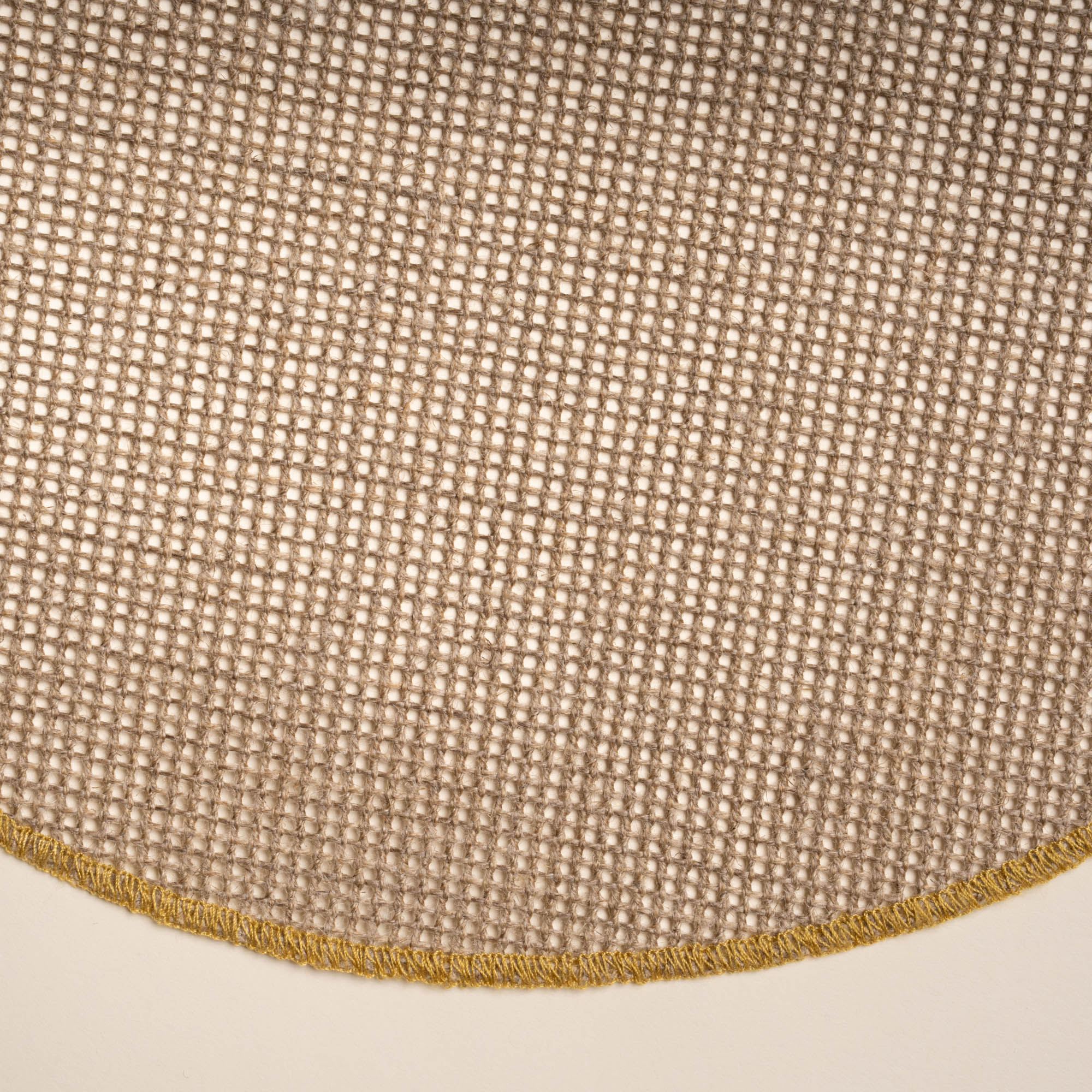 Closeup of a round woven placemat in a natural color with a sewn ochre rim on a beige background