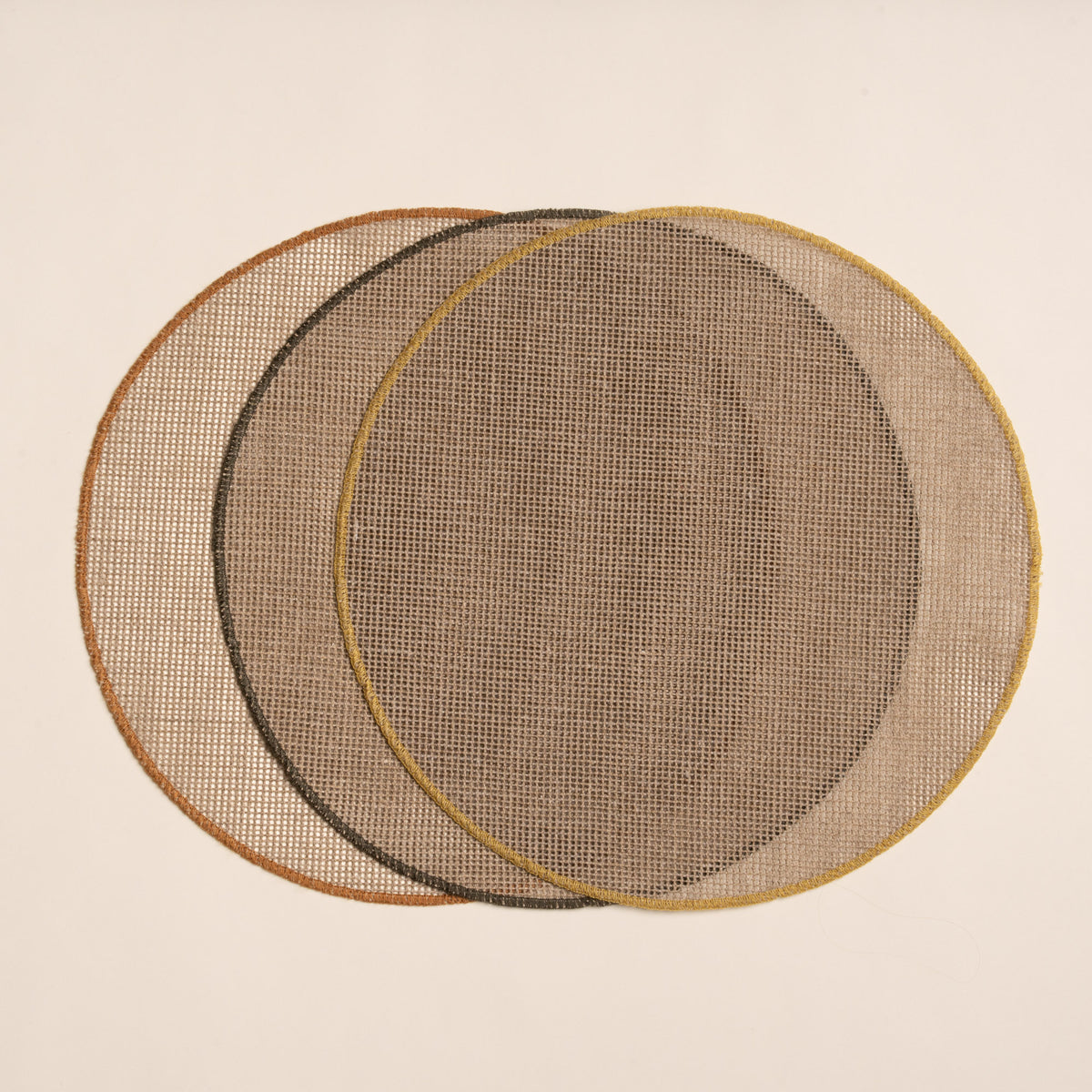 Three round woven placemats in a natural color with a sewn rust, ochre and olive green rims on a beige background