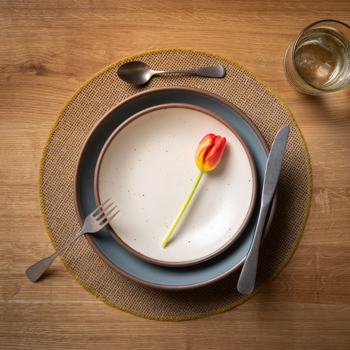 Dining table setting with plates, cutlery, and a tulip with a round woven placemat with a sewn ochre rim on a wooden surface