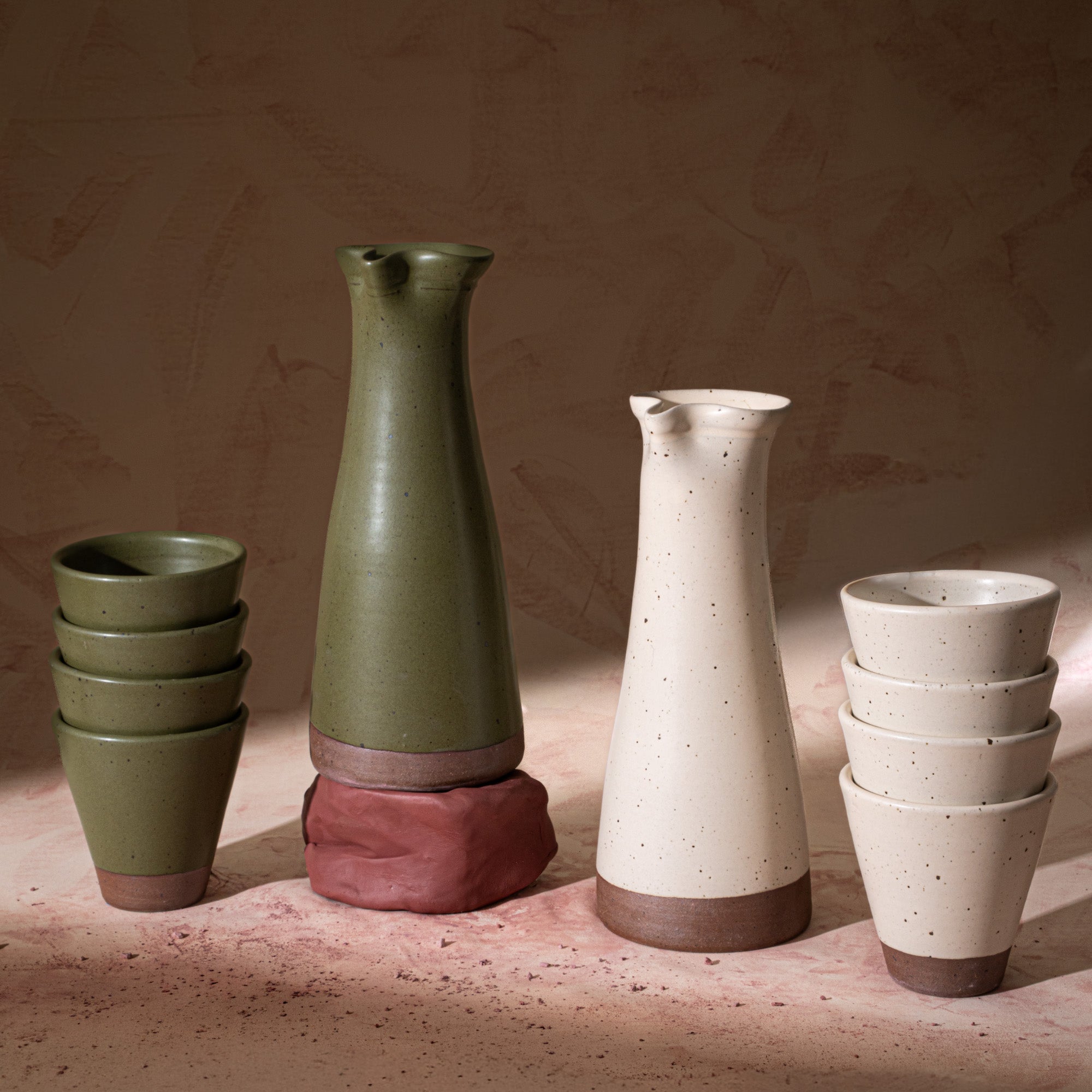 2 tall ceramic carafe and 2 stacsk of 4 tapered cups in an olive green and panna cotta colors, featuring iron speckles and unglazed bases, with some raw clay.