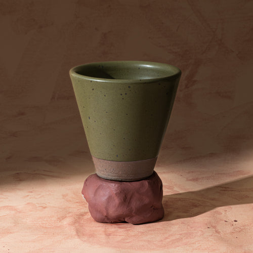 A tapered cup in an olive green color with iron speckles and unglazed base, sitting on raw clay.
