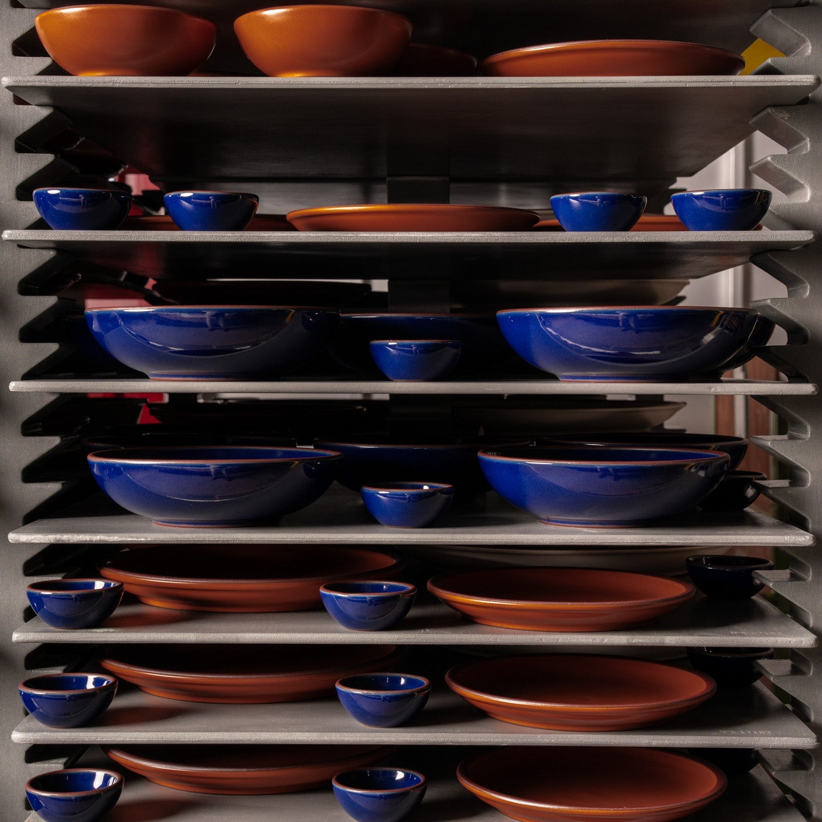 A kiln cart in the factory with glossy dark blue and earthy orange ceramic bowls and plates in various sizes.