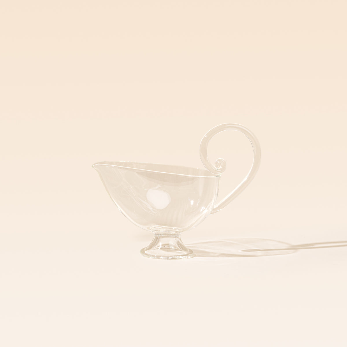 A clear glass sauce boat with a delicate curled handle, pointed pouring spout, and small pedestal foot, photographed on a warm cream background.