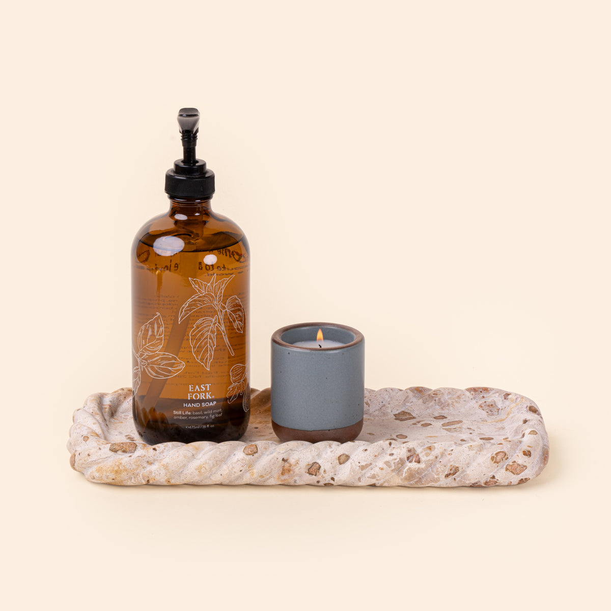 Hand Soap and small lit candle on a rectangular cream and brown speckled stone tray with a shallow recessed center and irregular scalloped edges.