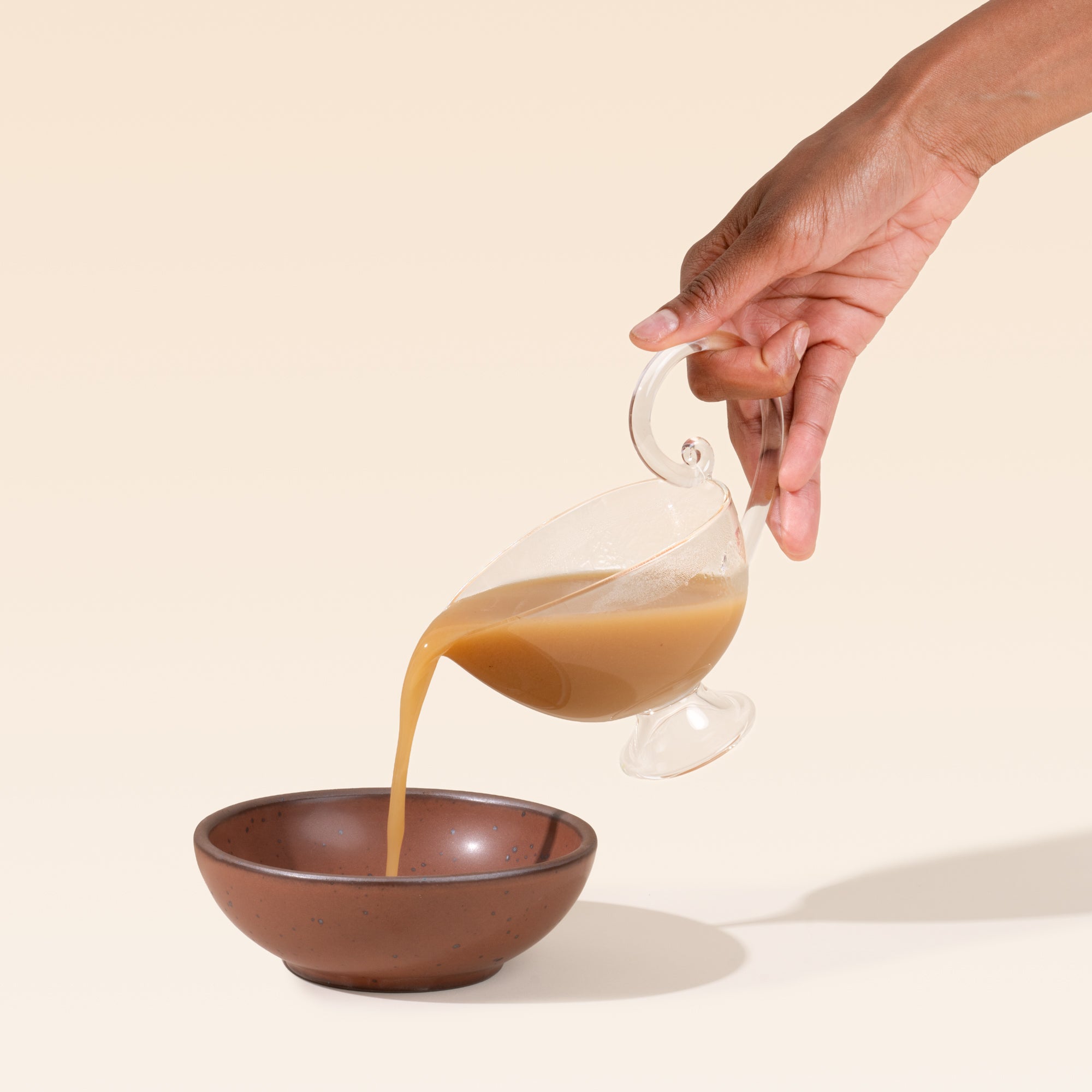 A hand holds a  clear glass gravy or sauce boat with a delicate curled handle, pointed pouring spout, and small pedestal foot, pouring sauce into a bowl.