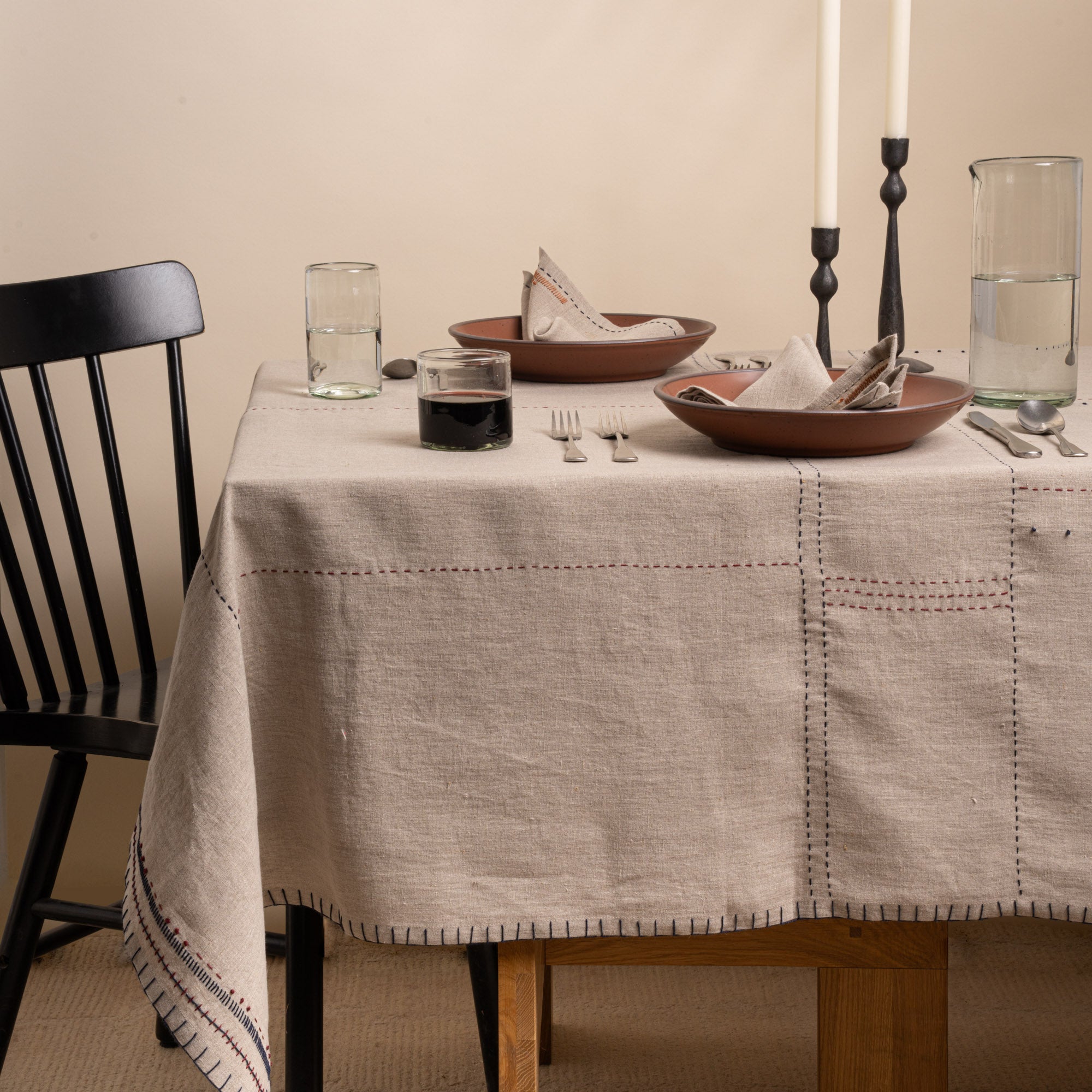 Natural linen tablecloth with hand-stitched navy and rust details, styled with warm ceramics, glassware, and black candlesticks.
