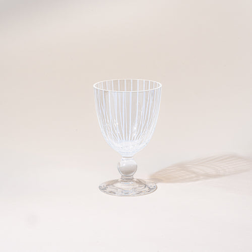 A clear stemmed goblet with a subtle striped pattern, photographed empty against a warm neutral background with a soft cast shadow.