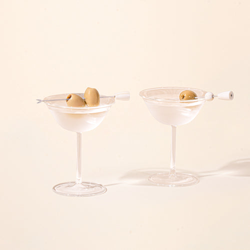 Two clear martini coupe glasses with cocktails, each garnished with green olives on cocktail picks.