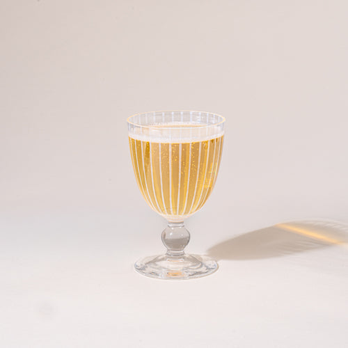 A clear stemmed goblet with a subtle polka-dot pattern, photographed against a warm neutral background with a soft cast shadow, filled with a sparkling drink.