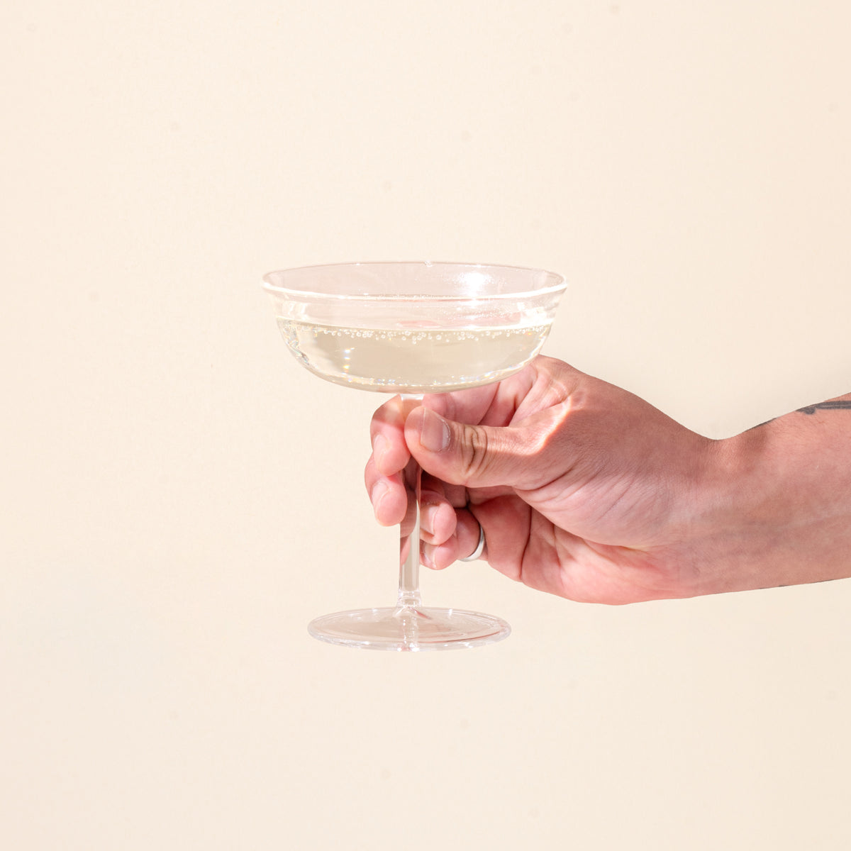 A hand holds a clear coupe glasses filled with champagne on a light background.